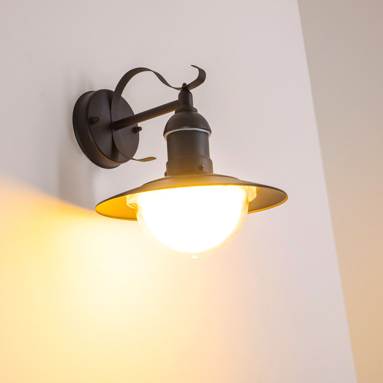 Eine dunkel gefärbte Wandleuchte mit leuchtender Glühbirne. Die Lampe befindet sich vor einer weißen Wand. Das Licht wirft einen warmen Schein.