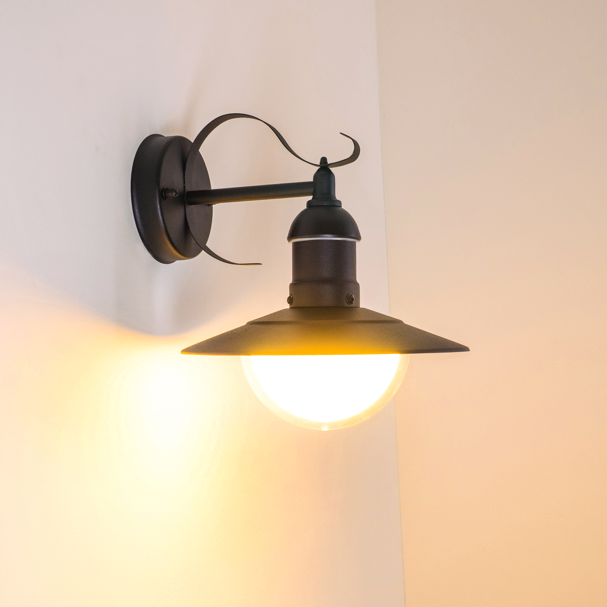 Eine schwarze Wandlampe mit nach unten gerichtetem Schirm, die warmes Licht ausstrahlt. Die Lampe ist an einer hellbeigen Wand angebracht.