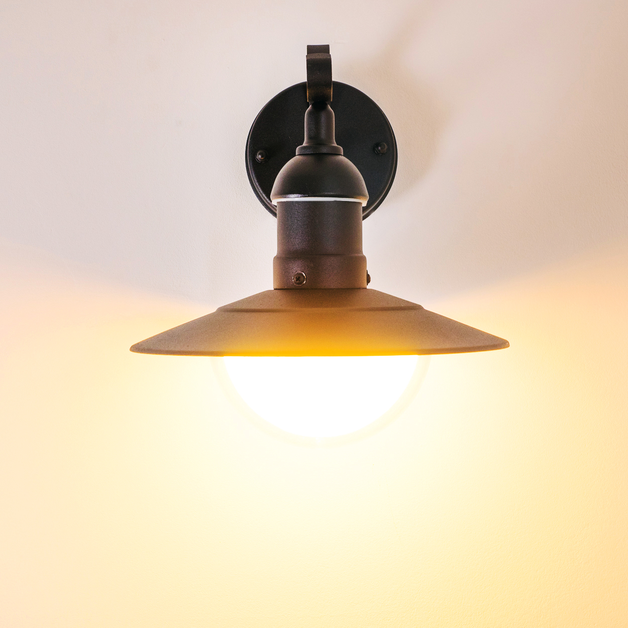 Eine schwarze Wandleuchte mit nach unten gerichtetem Schirm ist beleuchtet und wirft einen warmen Schein auf die Wand. Der Hintergrund ist cremefarben.
