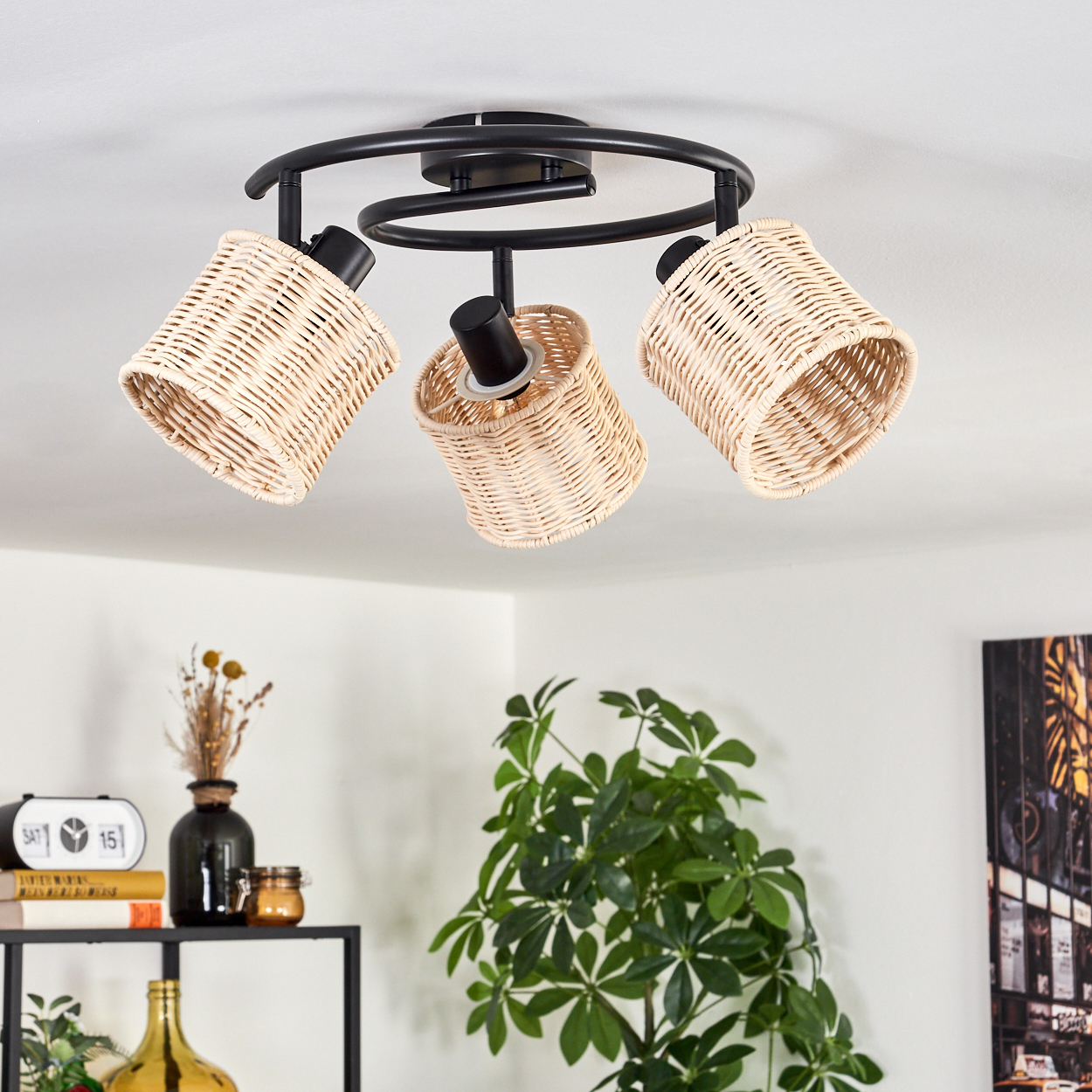 Eine Deckenlampe mit drei Korblampenschirmen hängt. Darunter sind ein Regal mit Dekoration und eine grüne Pflanze zu sehen.