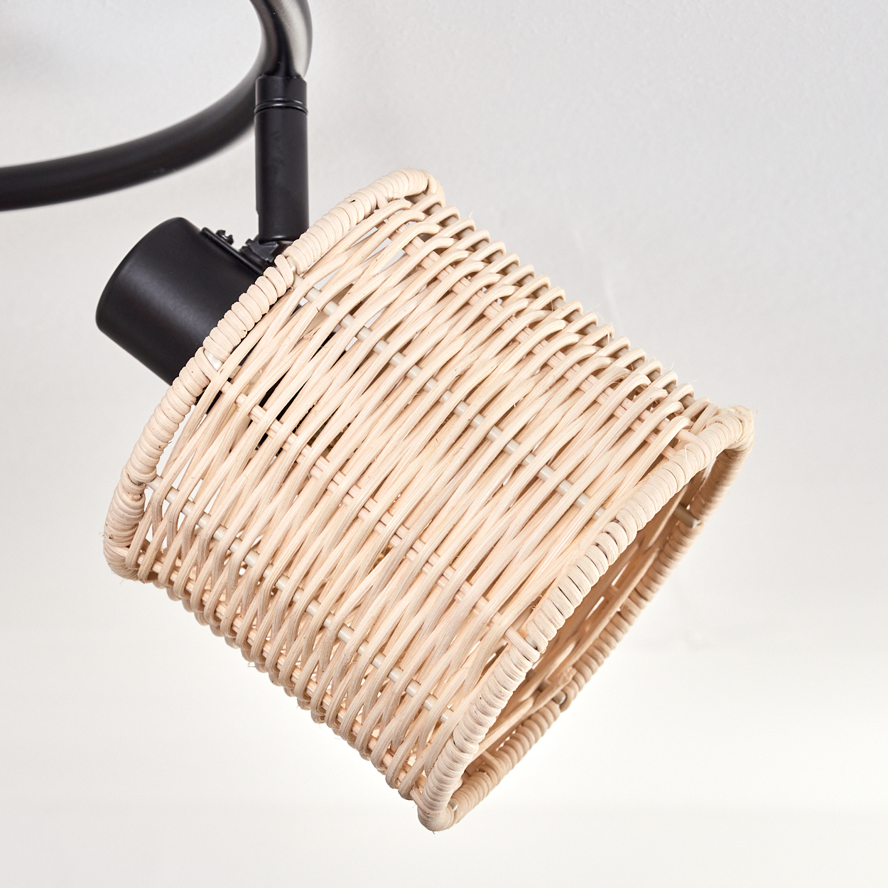 Ein Lampenschirm aus Korbgeflecht hängt an einer schwarzen Halterung vor weißem Hintergrund. Der Schirm ist beige und zylindrisch.