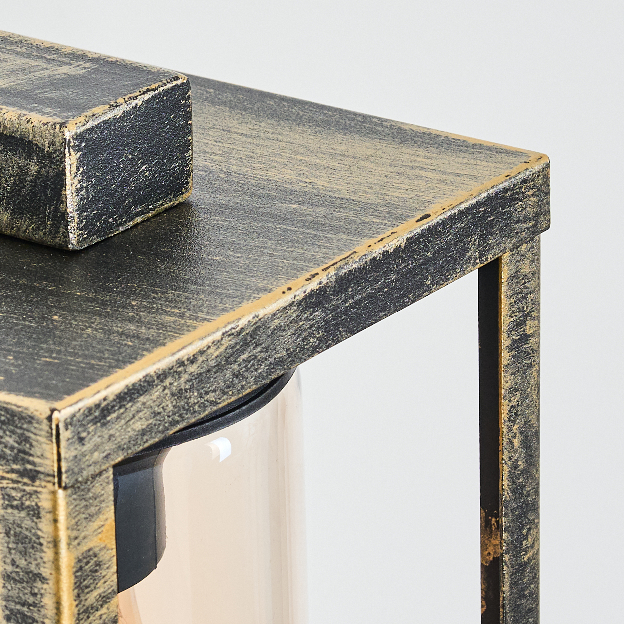 Nahaufnahme einer schwarz-goldenen Metalllaterne. Eine rechteckige Oberseite und ein kleiner Block sind sichtbar. Darunter ein Glaszylinder.