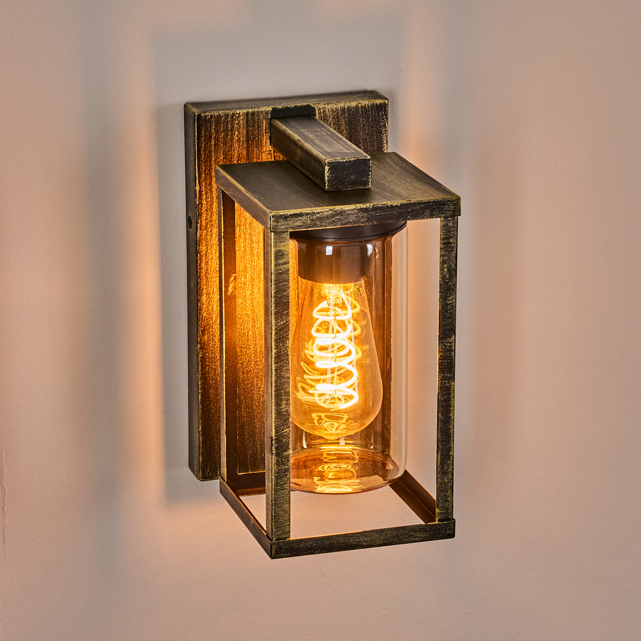 Eine Wandleuchte mit rechteckigem Rahmen und einer leuchtenden Glühbirne im Glas. Der Rahmen ist dunkelgold, an einer hellen Wand befestigt.