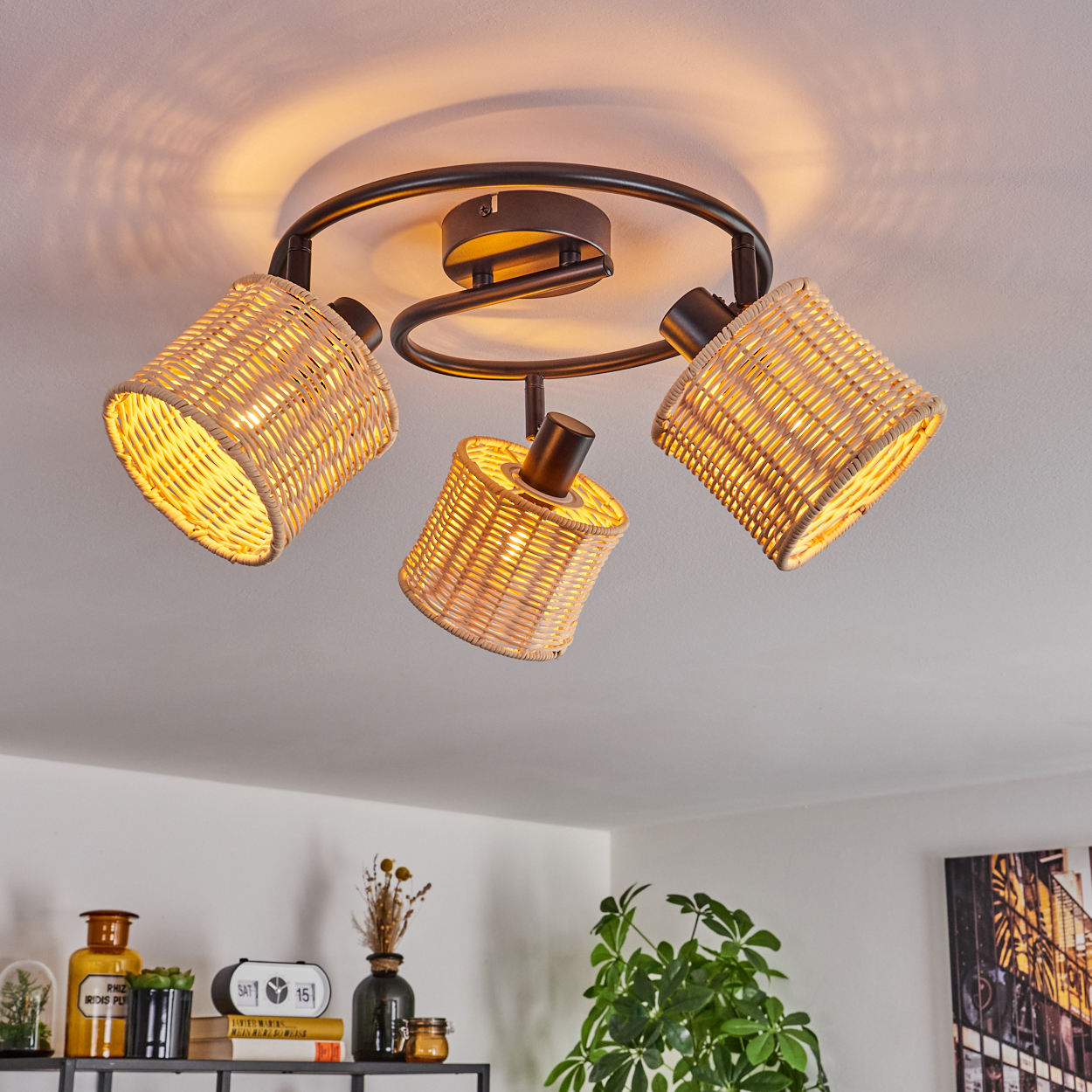 Eine Deckenleuchte mit drei Korblampenschirmen. Das Licht ist an. Im Hintergrund sind eine weiße Decke und einige Gegenstände auf einem Regal zu sehen.