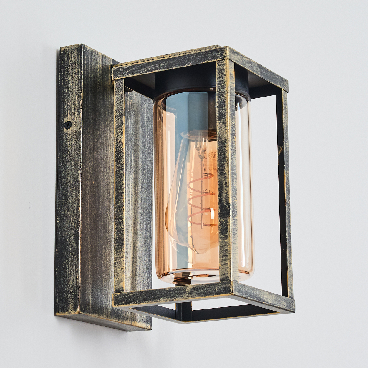 Eine Wandleuchte mit rechteckigem Metallrahmen, zylindrischem Glasschirm und sichtbarer Glühbirne vor weißer Wand.