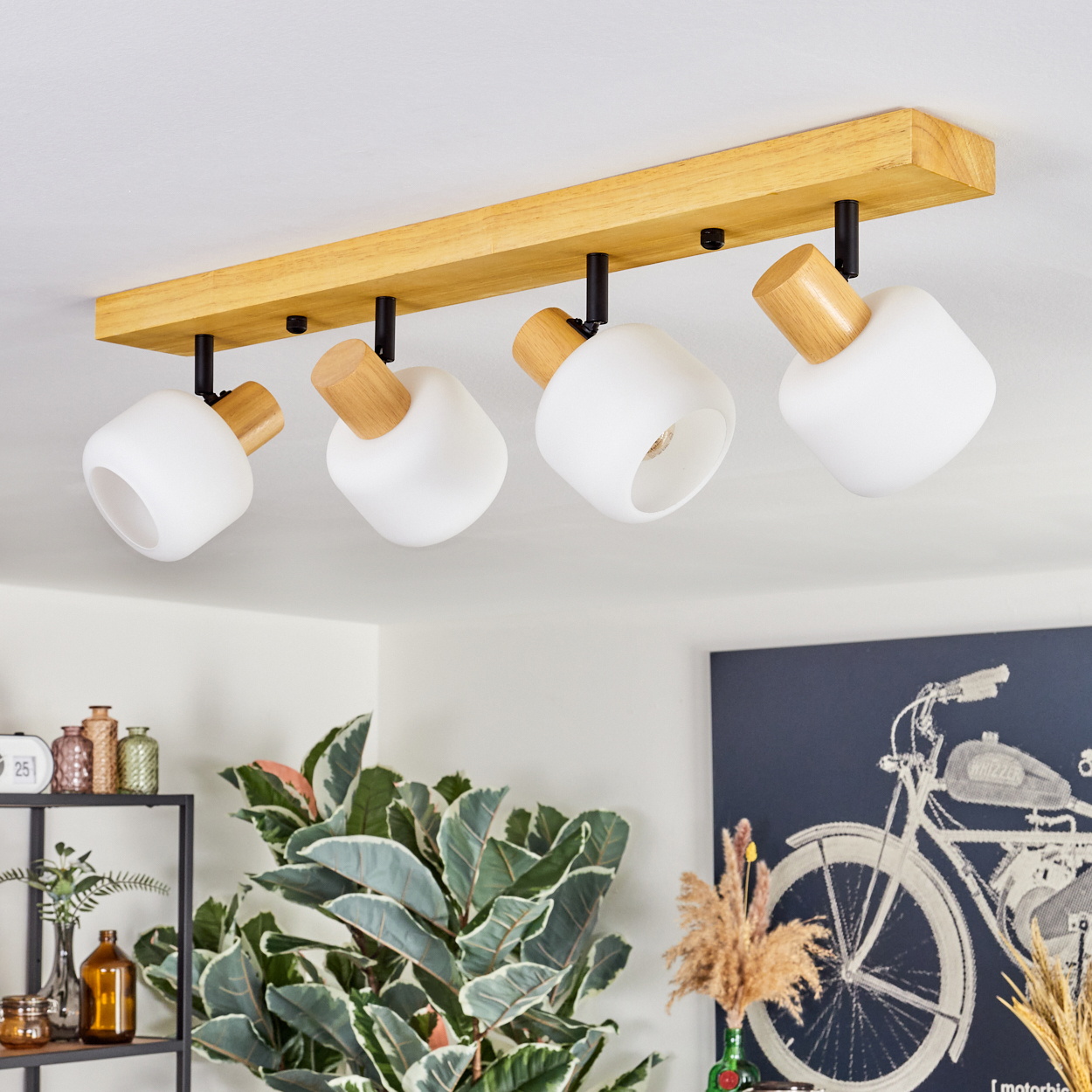 Eine Deckenleuchte mit vier weißen Lampen und Holzakzenten. Im Hintergrund sind eine Pflanze und ein Gemälde mit einem Fahrrad zu sehen.