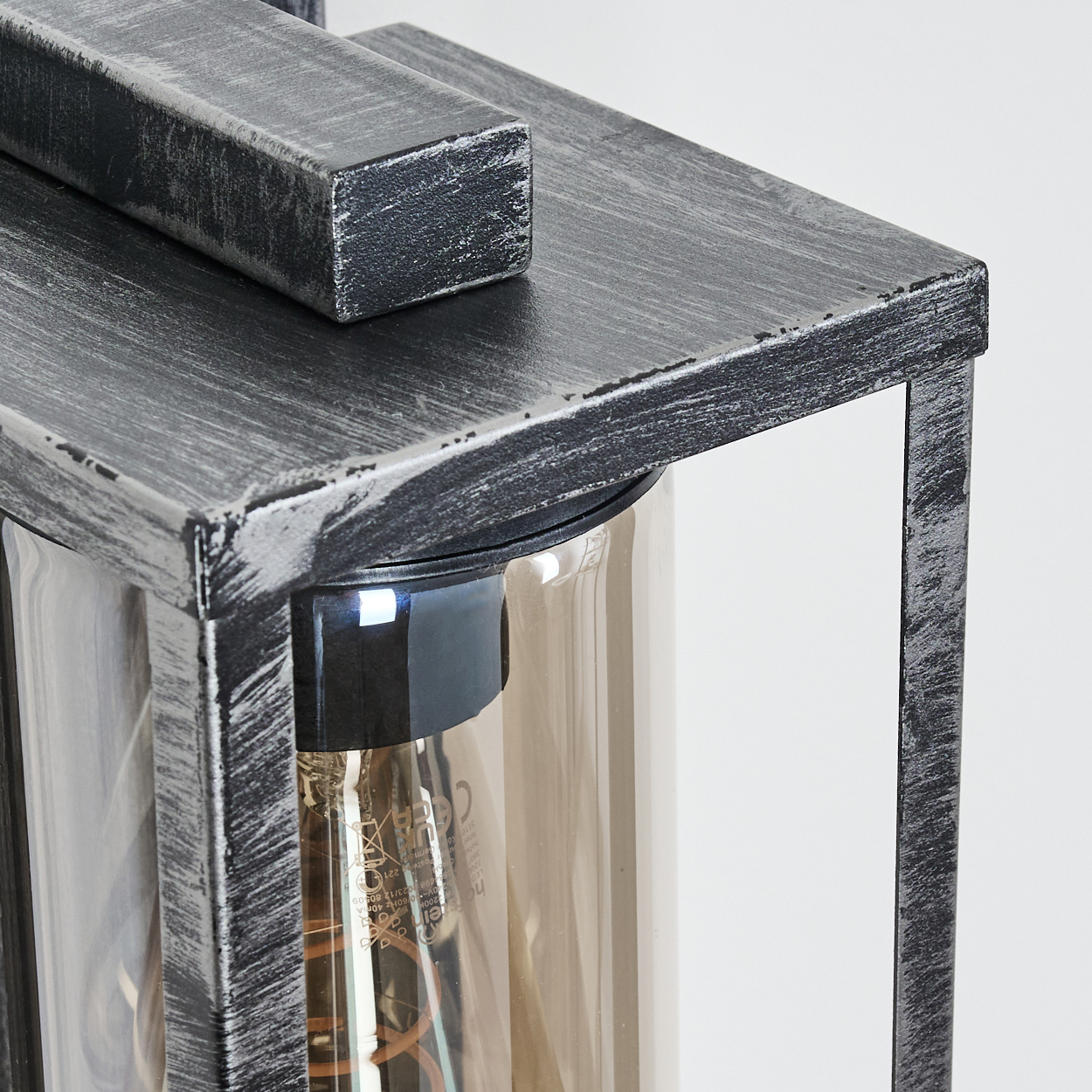 Nahaufnahme einer Metalllaterne mit klarer Glasumhüllung. Der Metallrahmen ist grau mit einem Used-Look, und die Glühbirne ist sichtbar.