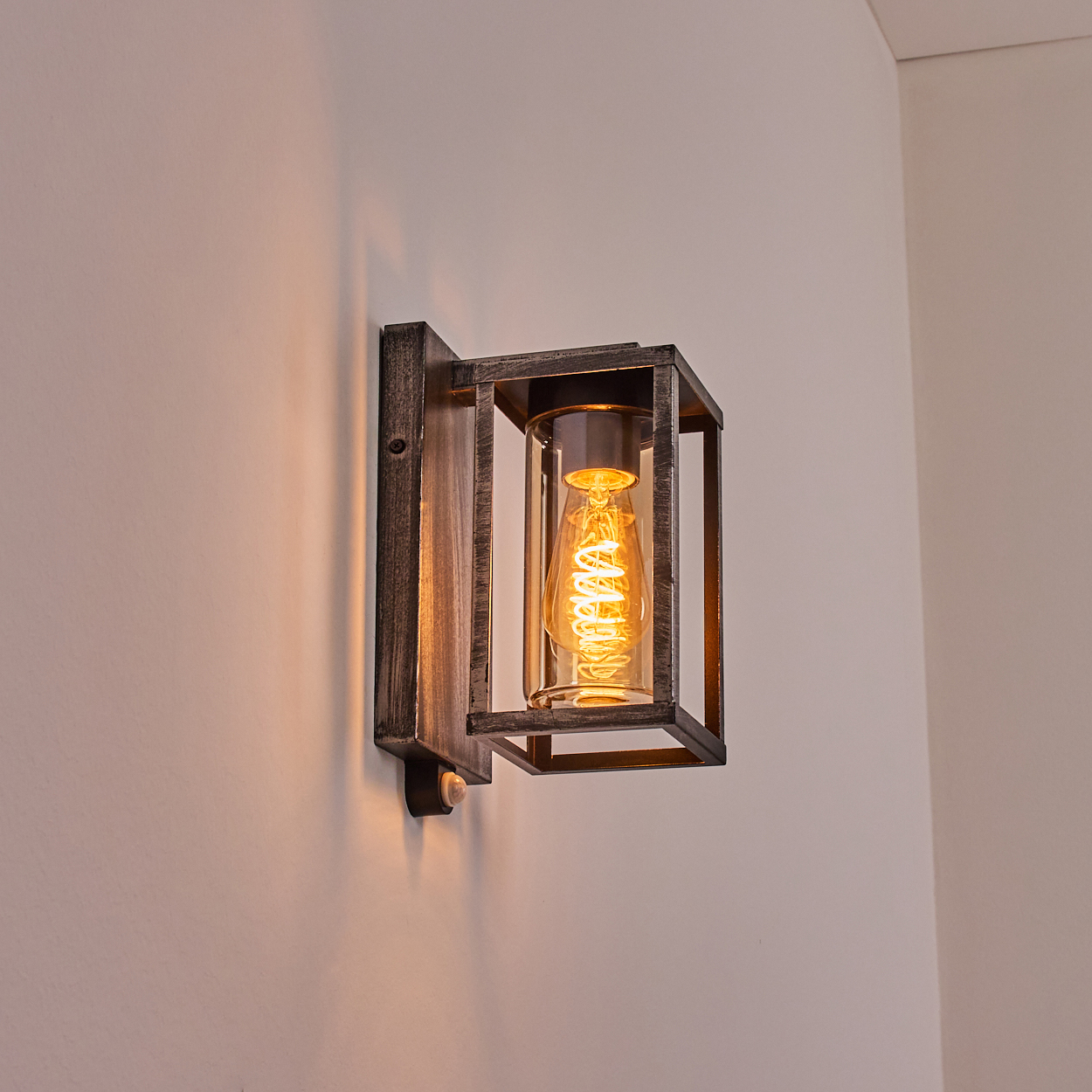 Eine Wandleuchte mit Vintage-Design. Sie hat einen rechteckigen Metallrahmen, der einen klaren Glaszylinder mit einer Spiralglühbirne umschließt.