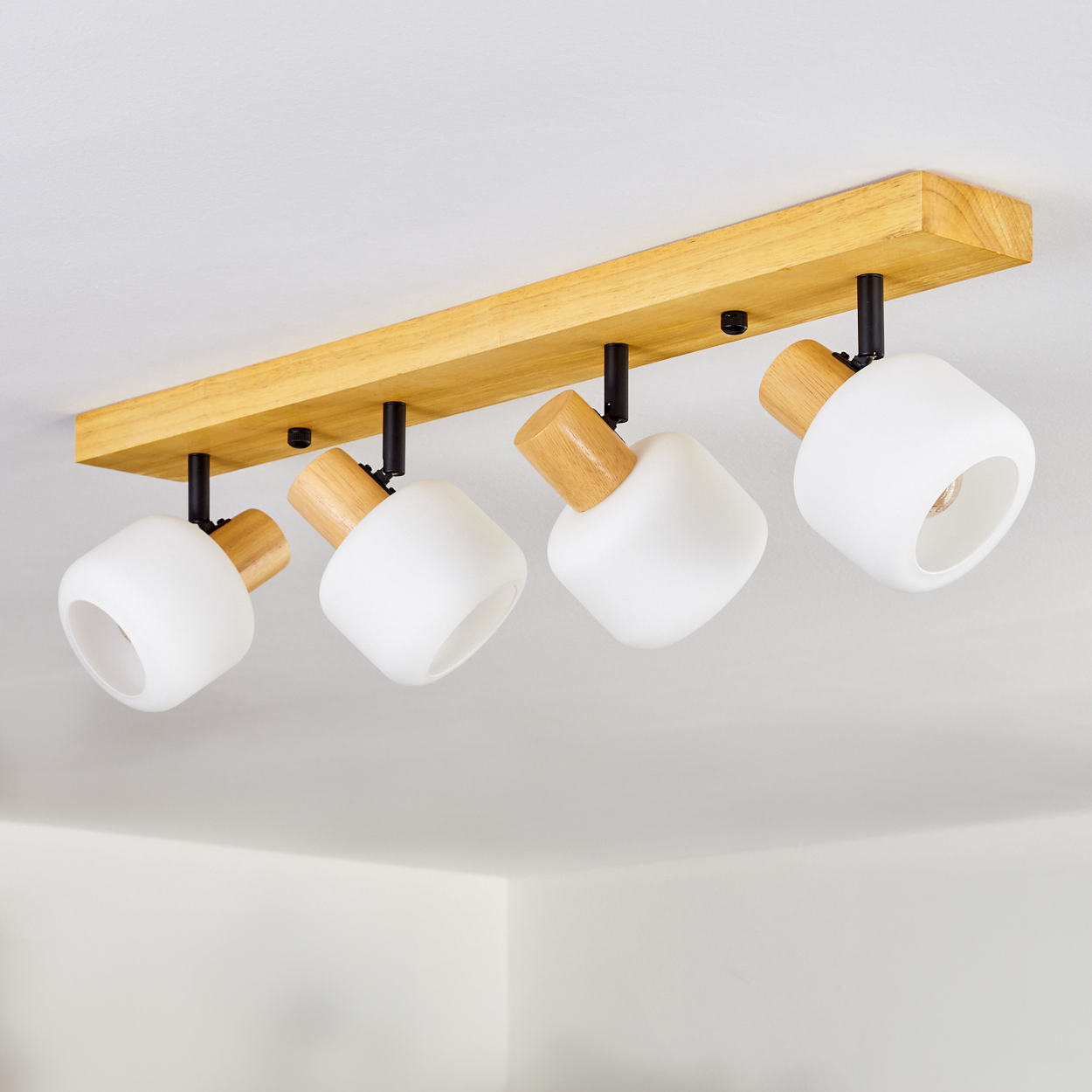 Eine Deckenleuchte mit vier weißen, kubusförmigen Lichtern, die an einem Holzbalken befestigt sind. Der Hintergrund ist eine weiße Decke.