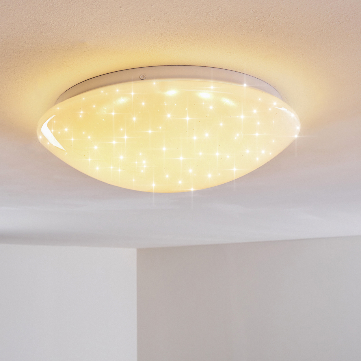 Eine ovale Deckenleuchte strahlt warmes Licht aus. Sie hat viele kleine, funkelnde Lichtpunkte und befindet sich an einer weißen Decke.