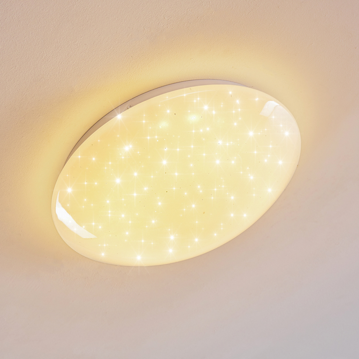 Eine ovale Deckenleuchte mit Sternenmuster. Das Licht ist weiß mit gelbem Schein vor einer weißen Decke.