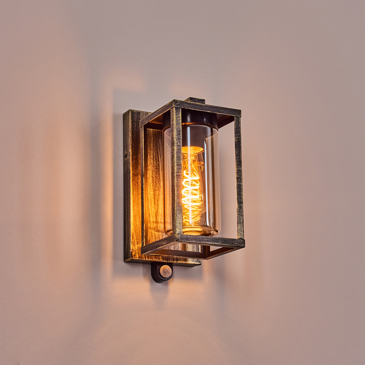 Wandleuchte mit rechteckigem Metallrahmen, Glasröhre und leuchtender Wendelbirne. An einer beigefarbenen Wand montiert.