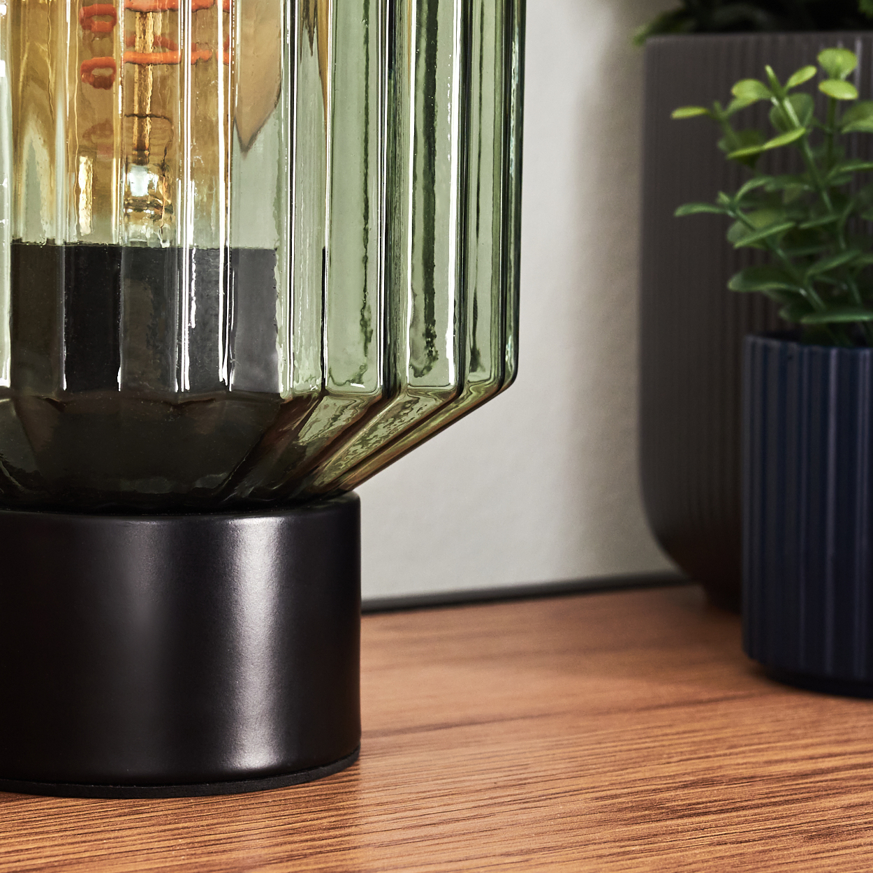 Nahaufnahme einer dekorativen Lampe mit grünem Glasschirm. Sie steht auf einer Holzoberfläche, im Hintergrund Pflanzen.