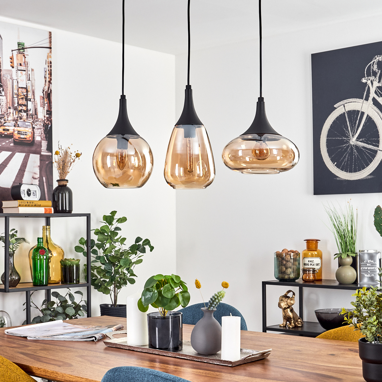 Drei Pendelleuchten über einem Holztisch. Die Dekoration umfasst Pflanzen, Vasen und Kunstwerke. Ein schwarzes Metallregal hält verschiedene Gegenstände.