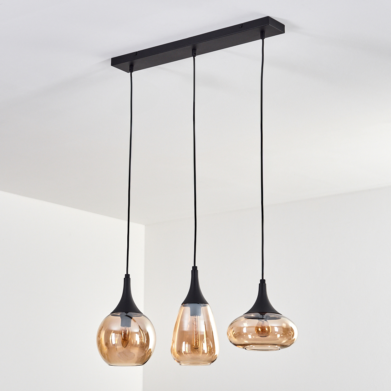 Drei Pendelleuchten mit bernsteinfarbenen Glasschirmen hängen an einer schwarzen Halterung an einer weißen Decke.