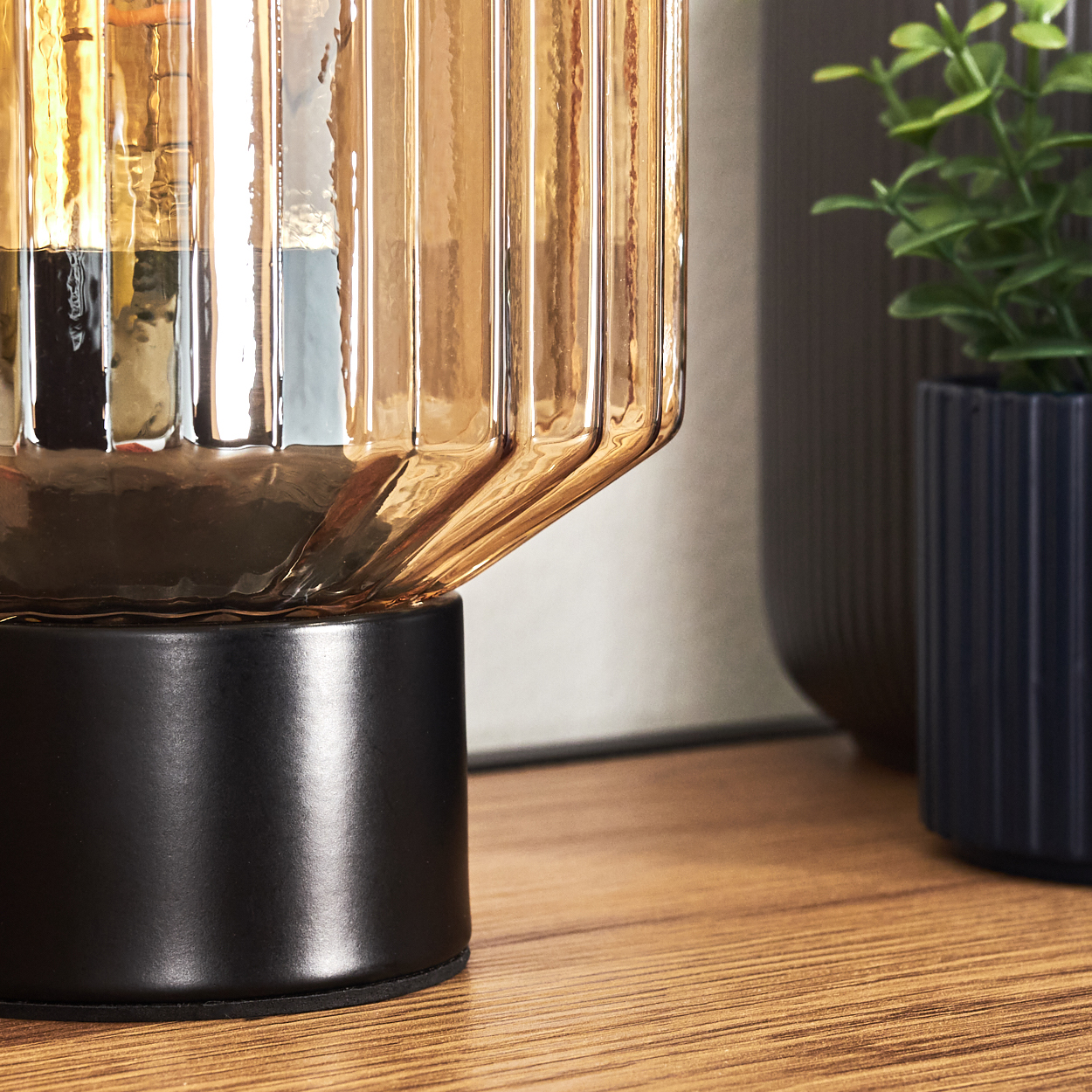 Nahaufnahme einer Lampe mit geripptem bernsteinfarbenem Glasschirm auf schwarzem Sockel, Holztisch und Pflanze in blauem Topf.