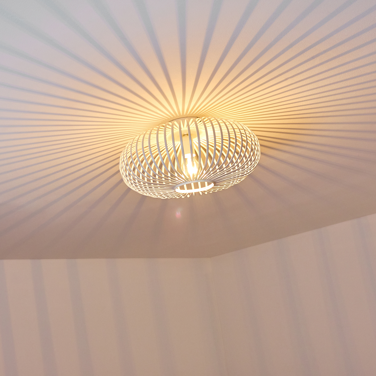Eine weiße, kugelförmige Lampe mit käfigartigem Design hängt von der Decke. Das Licht wirft Streifenschatten auf Decke und Wände.