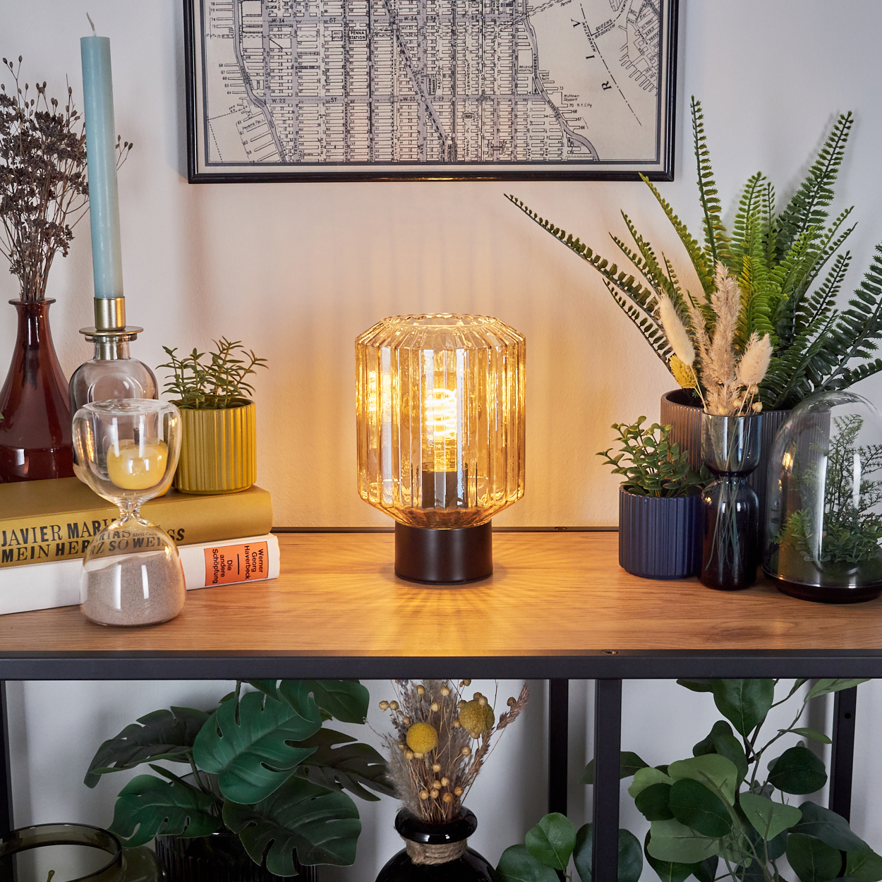 Eine Tischlampe mit Glasschirm, Bücher, Pflanzen und eine Karte. Auf dem Tisch befinden sich auch eine Kerze, eine Sanduhr und Vasen.