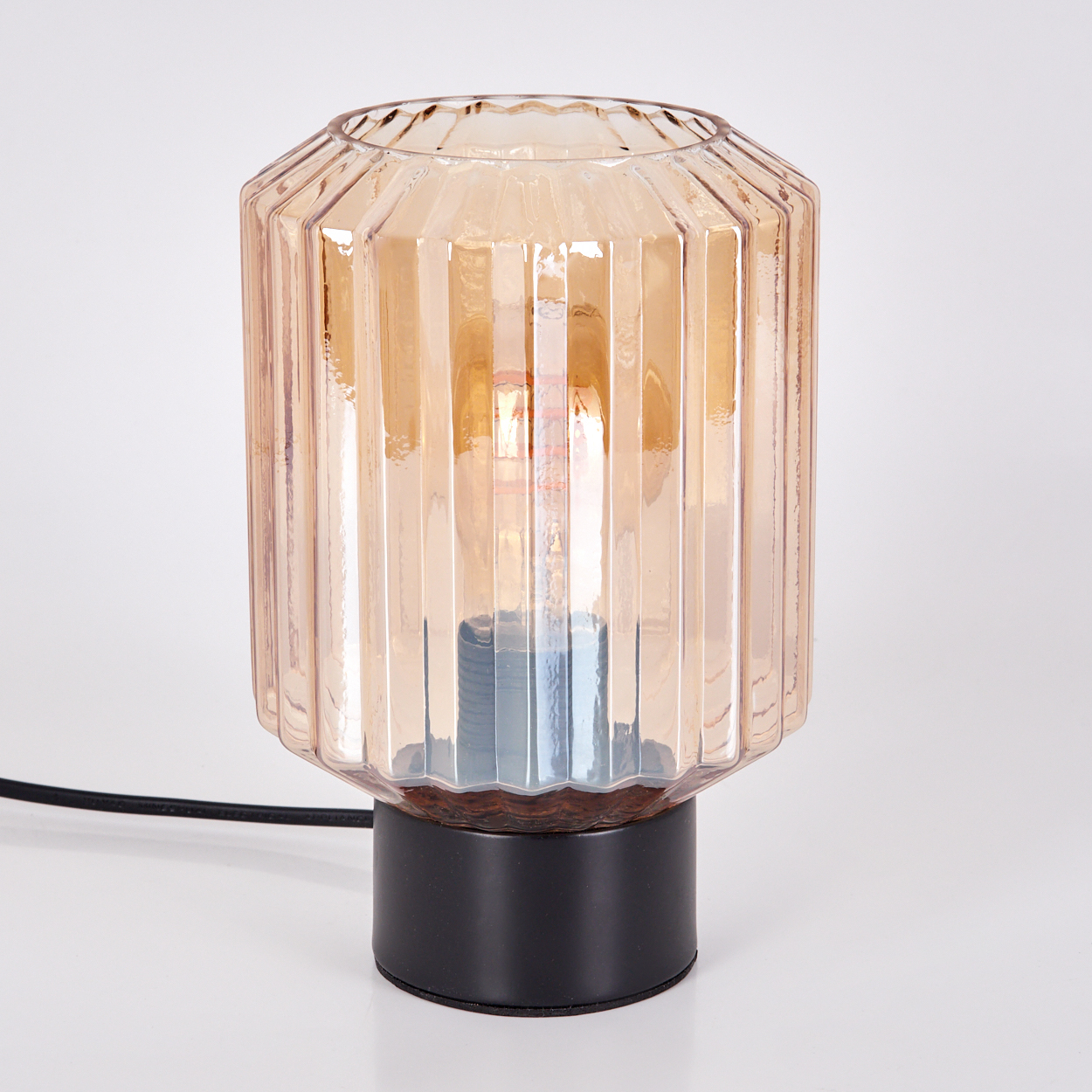Eine Glaslampe mit schwarzem Fuß. Das bernsteinfarbene Glas hat vertikale Rillen. Die Lampe steht auf weißem Untergrund.