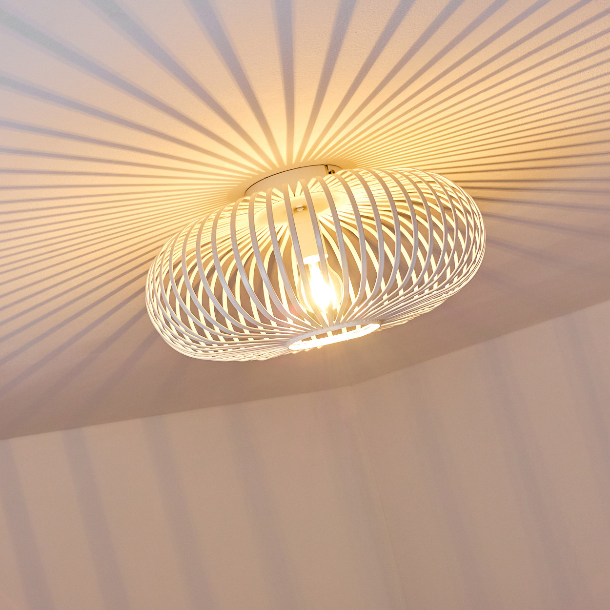 Eine weiße, käfigartige Lampe hängt von der Decke. Die Lampe wirft Schattenlinien auf die Decke. Das Licht ist an und hell.