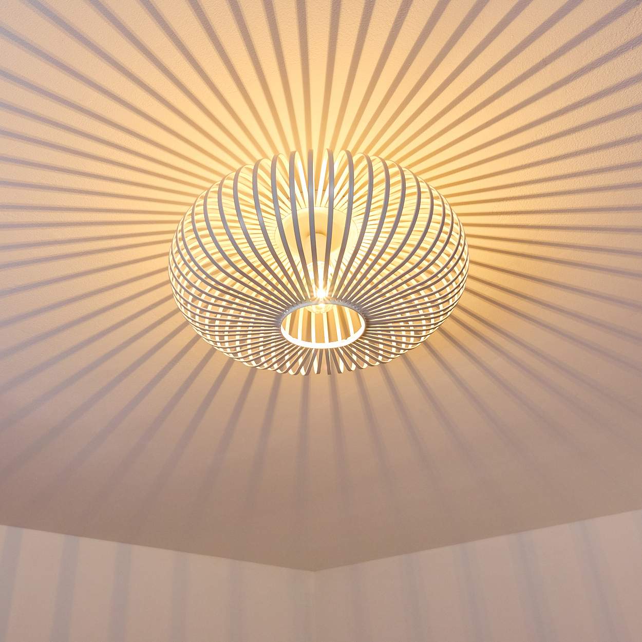 Eine moderne weiße Deckenlampe mit kreisförmigem Design, die radiale Schatten an die Decke wirft. Die Zimmerecken sind unten sichtbar.