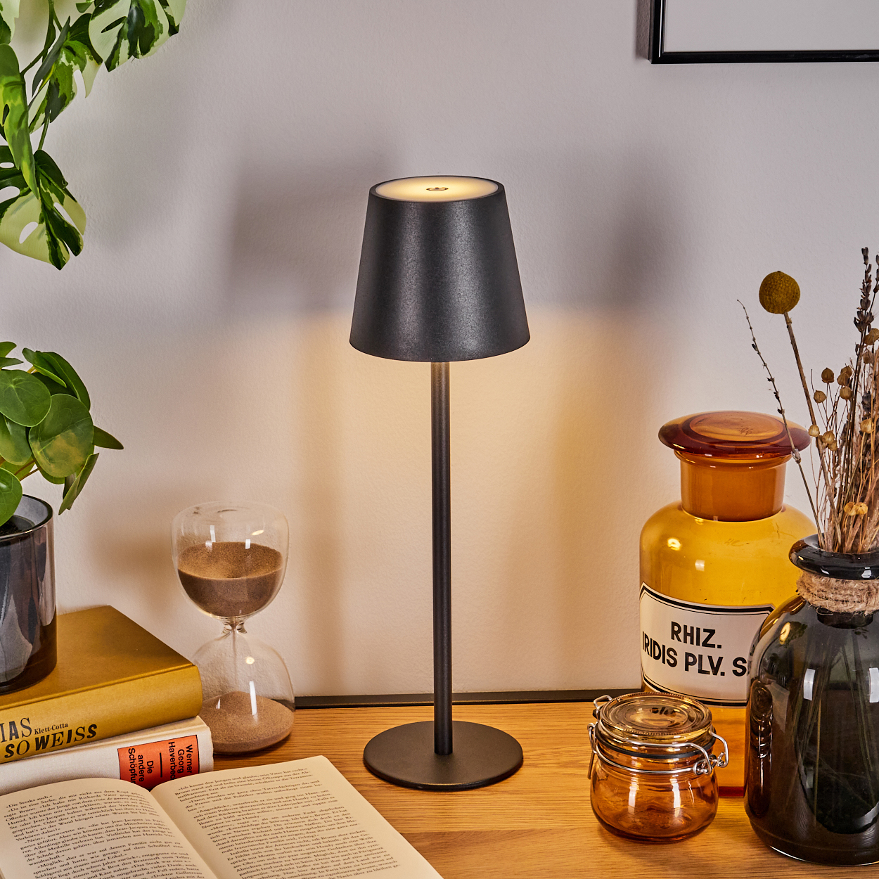 Eine schwarze Lampe auf einem Holztisch. Bücher, eine Sanduhr und Gläser stehen daneben. Grüne Pflanzen sind im Hintergrund.