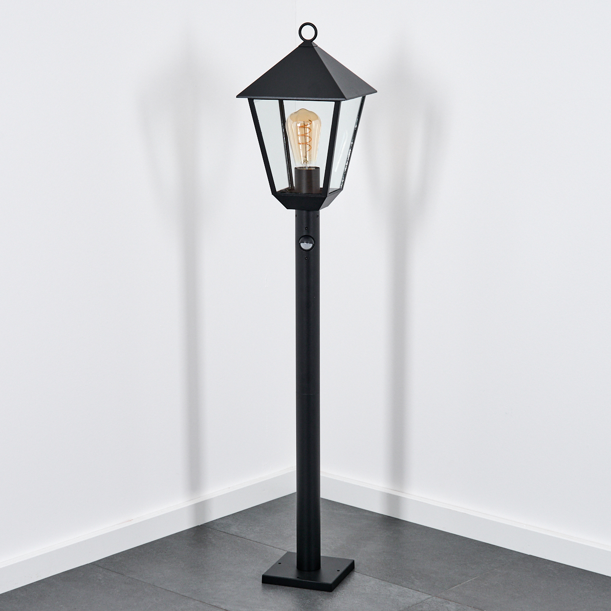 Eine schwarze Außenlampe mit Glasgehäuse und sichtbarer Glühbirne, die auf einem dunklen Fliesenboden steht, nahe einer weißen Wand.