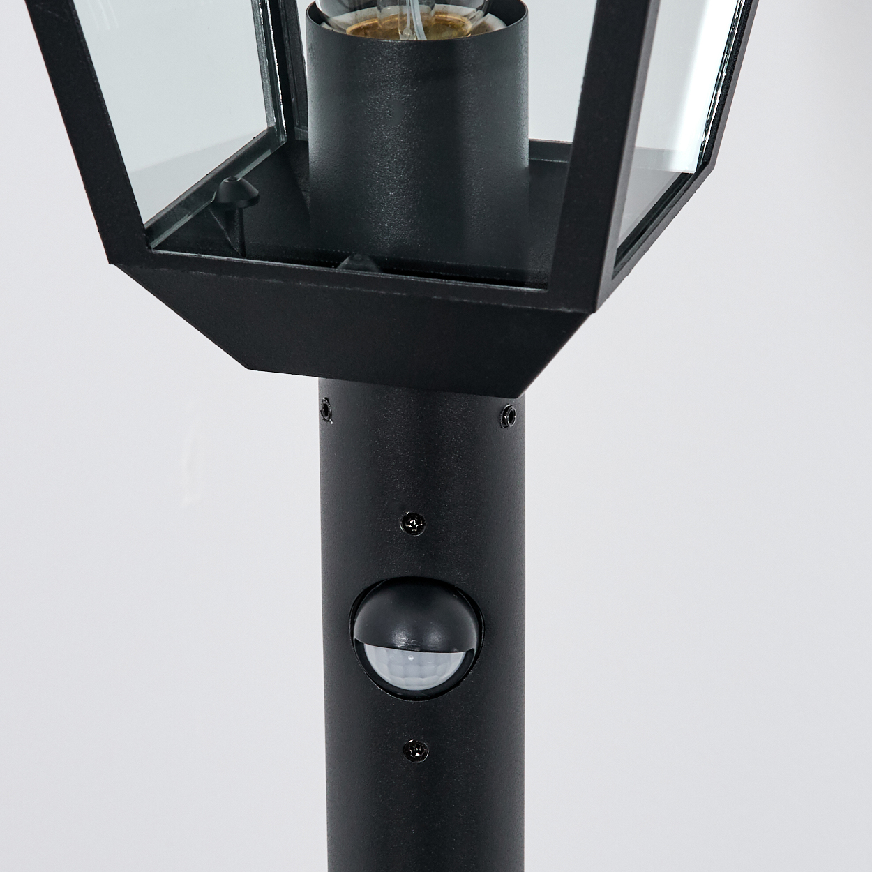 Nahaufnahme einer schwarzen Außenlampe. Sie hat einen transparenten Glasschirm, eine Glühbirne und einen Sensor. Der Hintergrund ist weiß.