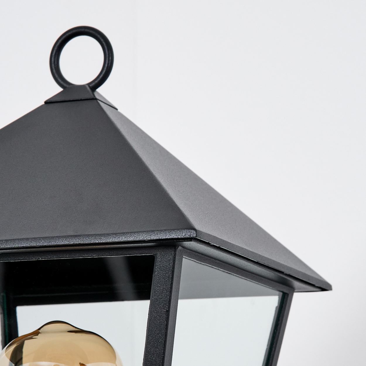 Nahaufnahme einer schwarzen Außenlampe. Oben befindet sich ein schwarzer Metallring. Die Lampe hat Glasscheiben und eine Glühbirne.