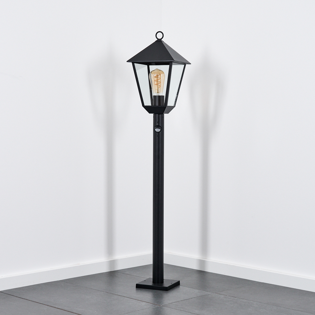 Eine schwarze Außenlampe steht in einer Ecke. Sie hat ein glasumschlossenes Licht und eine hohe, dünne Stange. Der Hintergrund ist weiß.