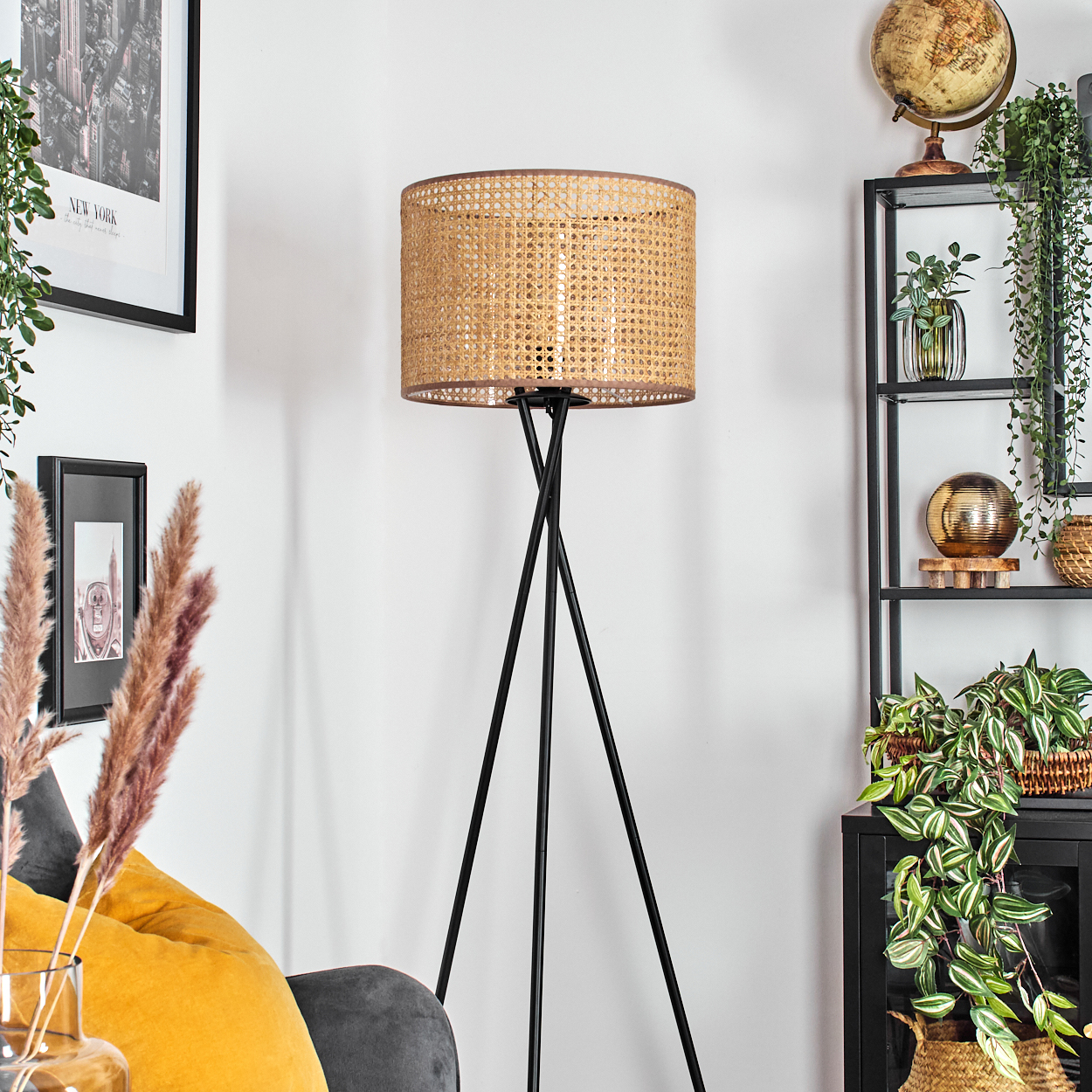Eine Stehlampe mit gewebtem Schirm steht in einem Raum mit Regal, Globus und Pflanzen. Ein Bild von New York hängt an der Wand.