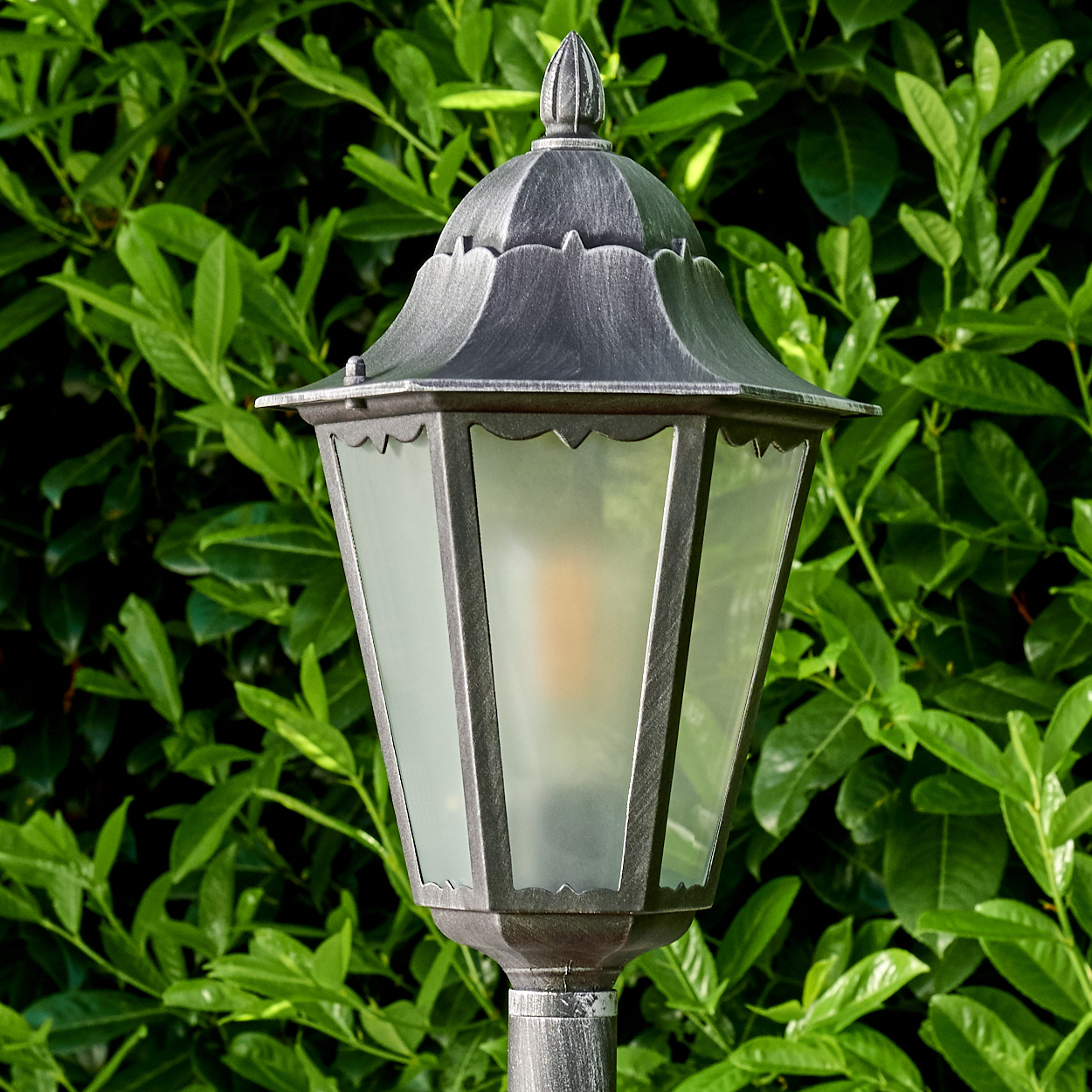 Eine metallisch-graue Außenlampe mit Glasscheiben steht vor einem Hintergrund aus üppigen grünen Blättern. Die Lampe leuchtet warm.