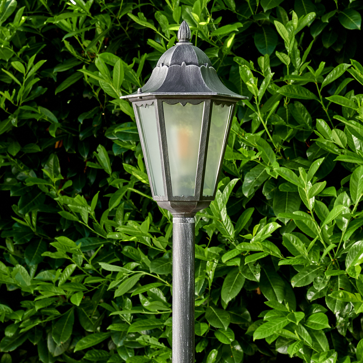 Eine graue Laterne auf einem Pfahl steht vor einer dichten grünen Hecke. Das Licht der Laterne ist schwach sichtbar.