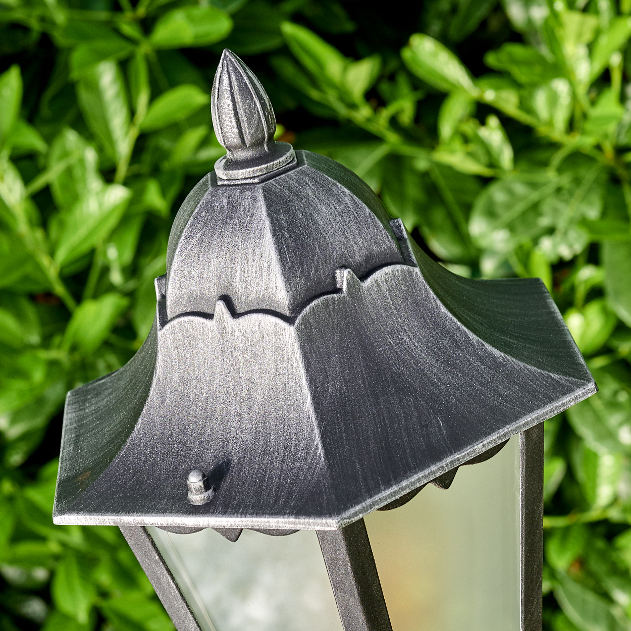 Nahaufnahme einer dekorativen Metalllampe mit grünem Blätterhintergrund, mit Fokus auf der Oberseite und Glasscheibe.