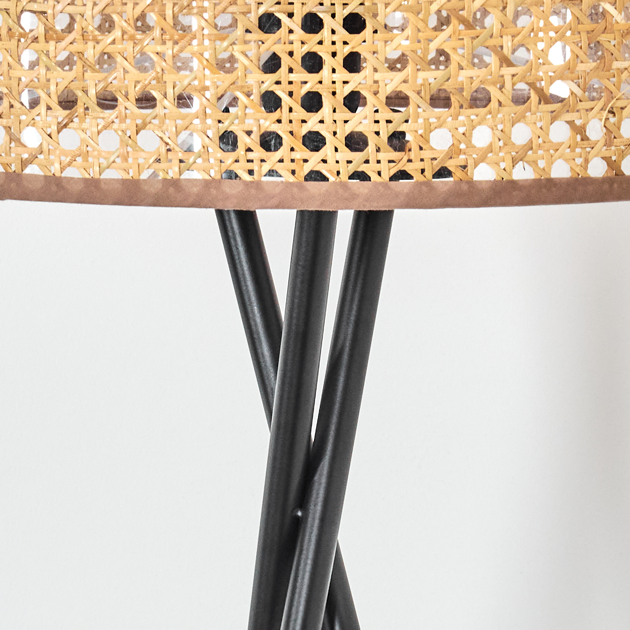 Nahaufnahme eines Lampenschirms aus geflochtenem Rattan mit dunklen Metallbeinen. Der Schirm hat eine braune Zierleiste. Der Hintergrund ist weiß.