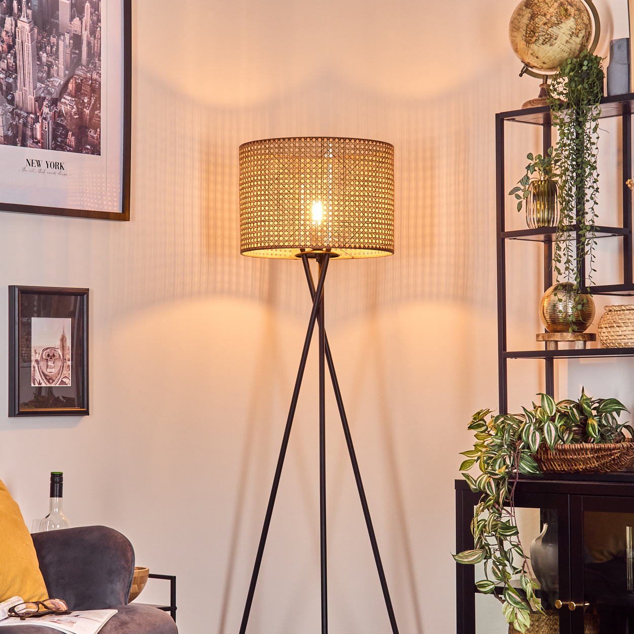 Eine beleuchtete gewebte Lampe steht in einem Raum. Ein schwarz gerahmtes New York-Bild hängt an der Wand. Rechts ein schwarzes Regal mit Pflanzen.