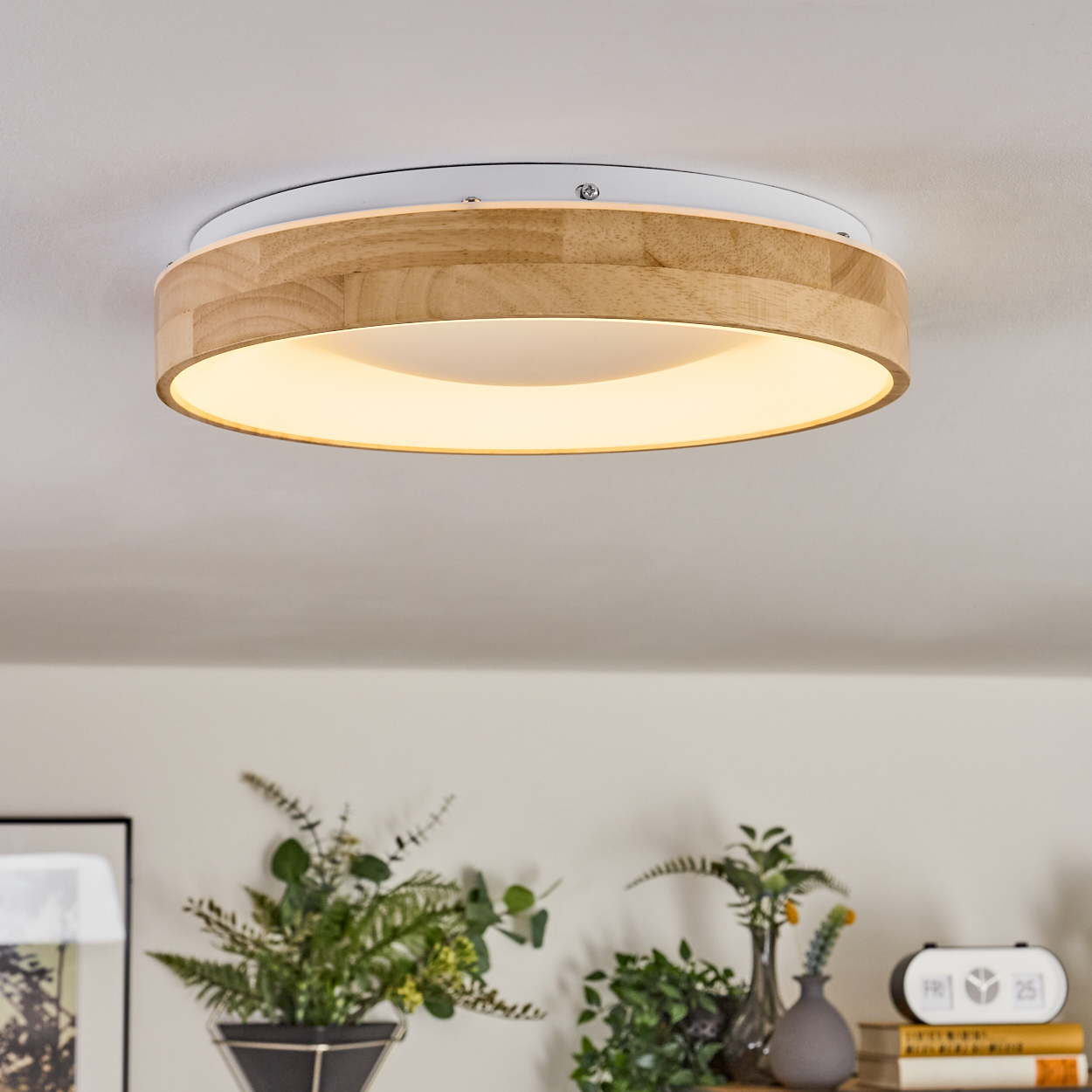 Eine runde Deckenlampe aus Holz mit weißem Sockel. Darunter Pflanzen in Vasen und ein Wecker auf einem Regal, alles in einem Raum.
