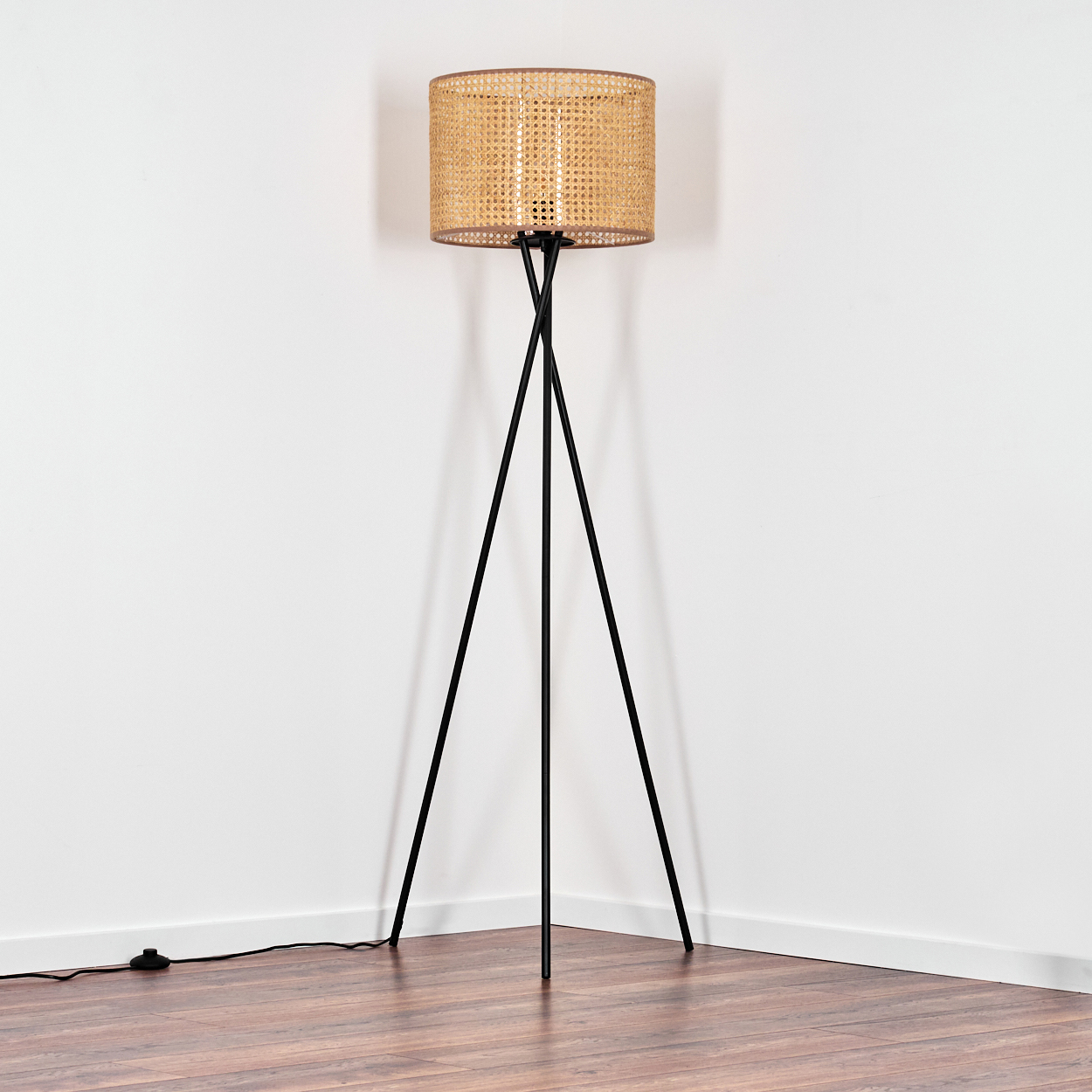 Eine Stehlampe mit gewebtem Lampenschirm steht in der Ecke eines Raumes mit weißen Wänden und Holzboden.