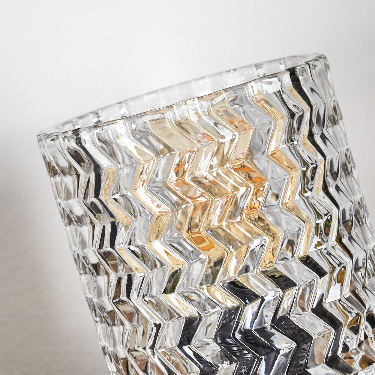 Eine klare Glasvase mit Zickzackmuster. Die Vase reflektiert Licht und erzeugt ein Spiel von Silber- und Goldtönen.