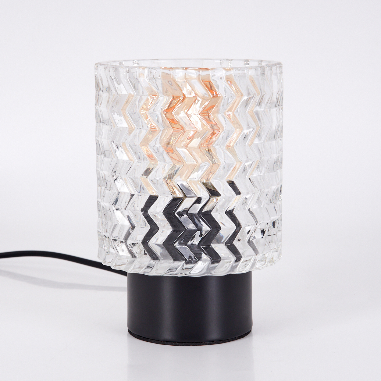 Eine klare Glaslampe mit schwarzem Fuß und Zickzackmuster. Das Licht im Inneren ist orange. Ein schwarzes Kabel ist angeschlossen.