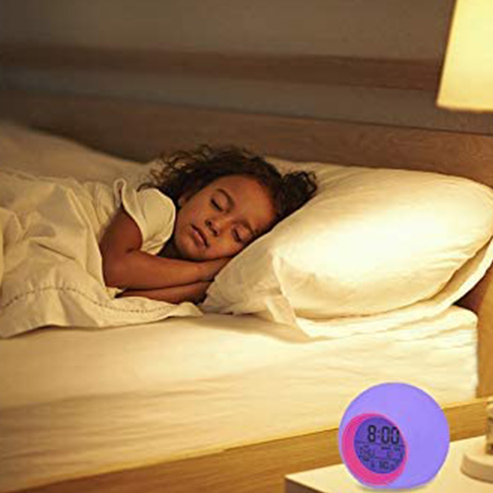 Ein junges Mädchen schläft im Bett mit weißem Kissen und Decke. Ein bunter Wecker steht daneben.