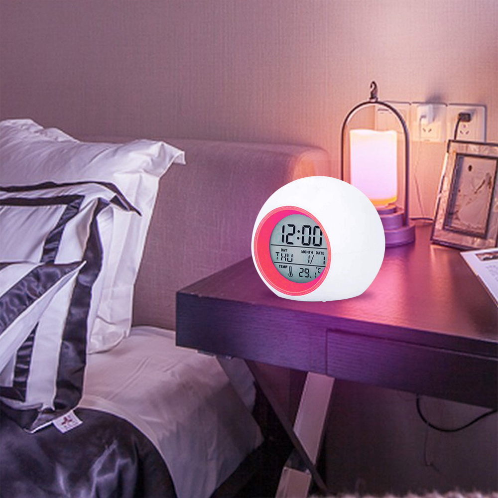 Ein weißer und rosafarbener digitaler Wecker zeigt Uhrzeit, Datum und Temperatur auf einem Nachttisch neben einem Bett an. Eine Lampe und ein Bilderrahmen sind ebenfalls sichtbar.