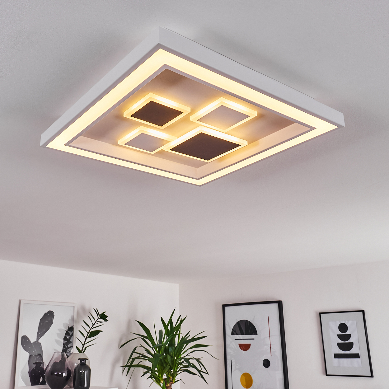 Moderne Deckenleuchte mit quadratischem Design, die warmes Licht ausstrahlt. Darunter dekorieren Pflanzen und gerahmte Kunstwerke eine weiße Wand.