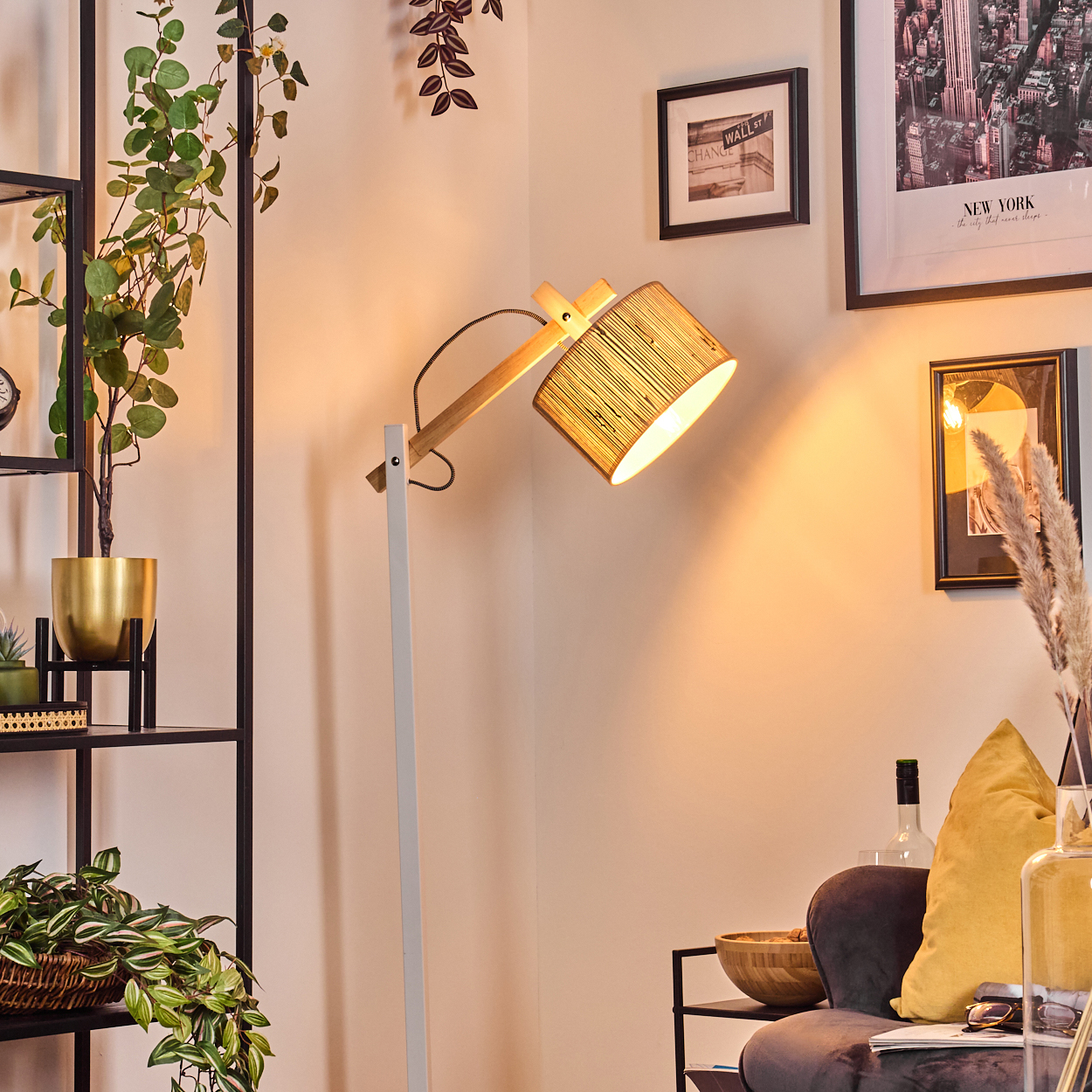 Eine beleuchtete Stehlampe mit Bambusschirm. Ein schwarzes Regal mit Pflanzen und einem goldenen Topf. Bilder an der Wand und ein gelbes Kissen.