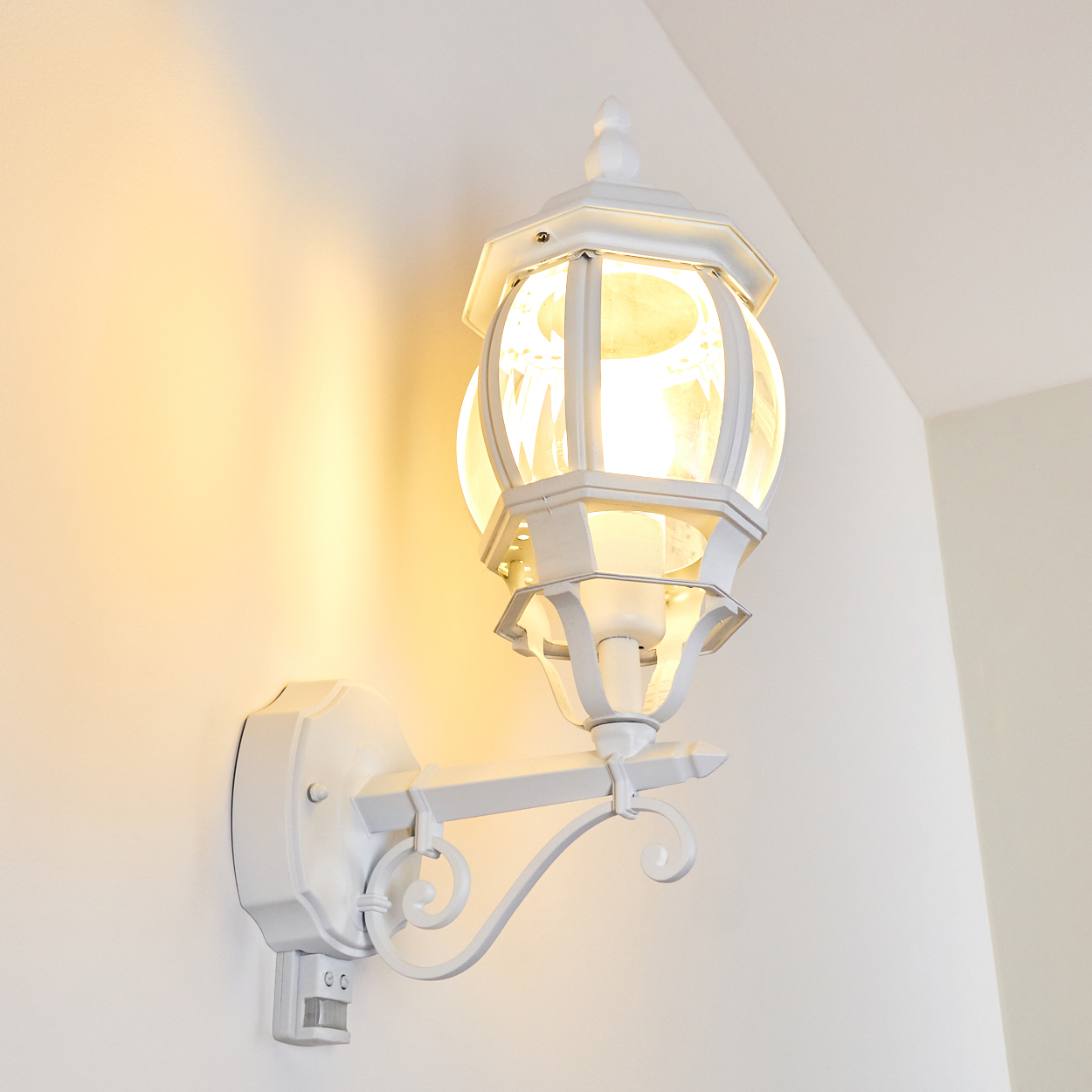 Weiße Außenwandleuchte mit Glaskugel, an einer weißen Wand montiert. Die Lampe ist beleuchtet.