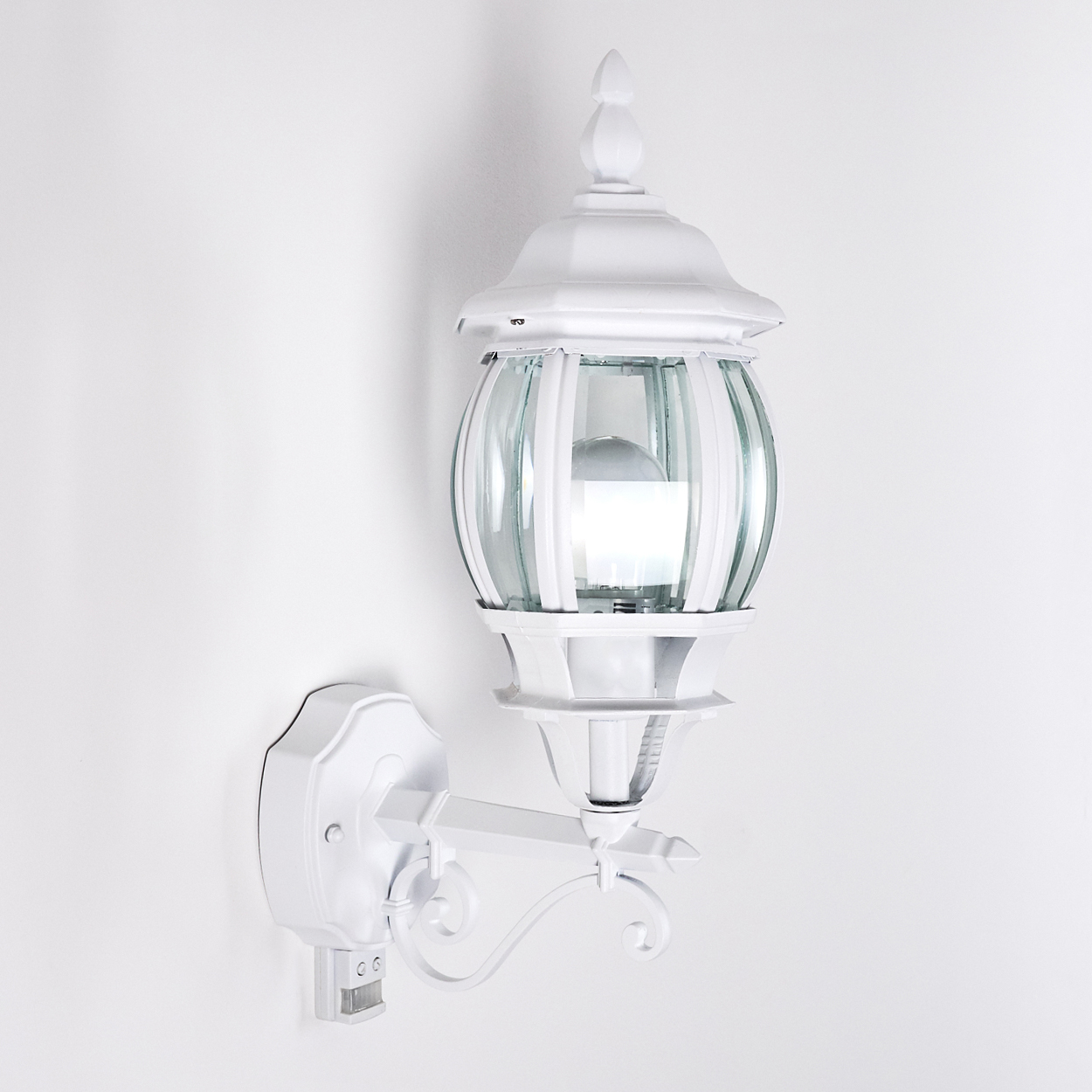Eine weiße, verzierte Außenlampe ist an einer weißen Wand montiert. Die Lampe hat Glasplatten und eine dekorative Spitze.