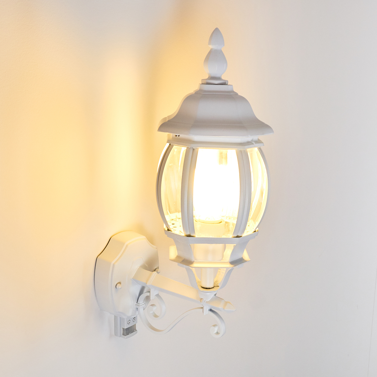 Eine weiße Außenwandleuchte ist an einer weißen Wand montiert. Die Lampe hat eine Glaskugel und eine dekorative Spitze.