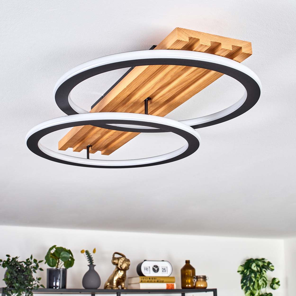 Moderne Deckenleuchte mit zwei schwarz-weißen Ringen, Holzelementen und einem Regal mit Dekoration in einem Raum.