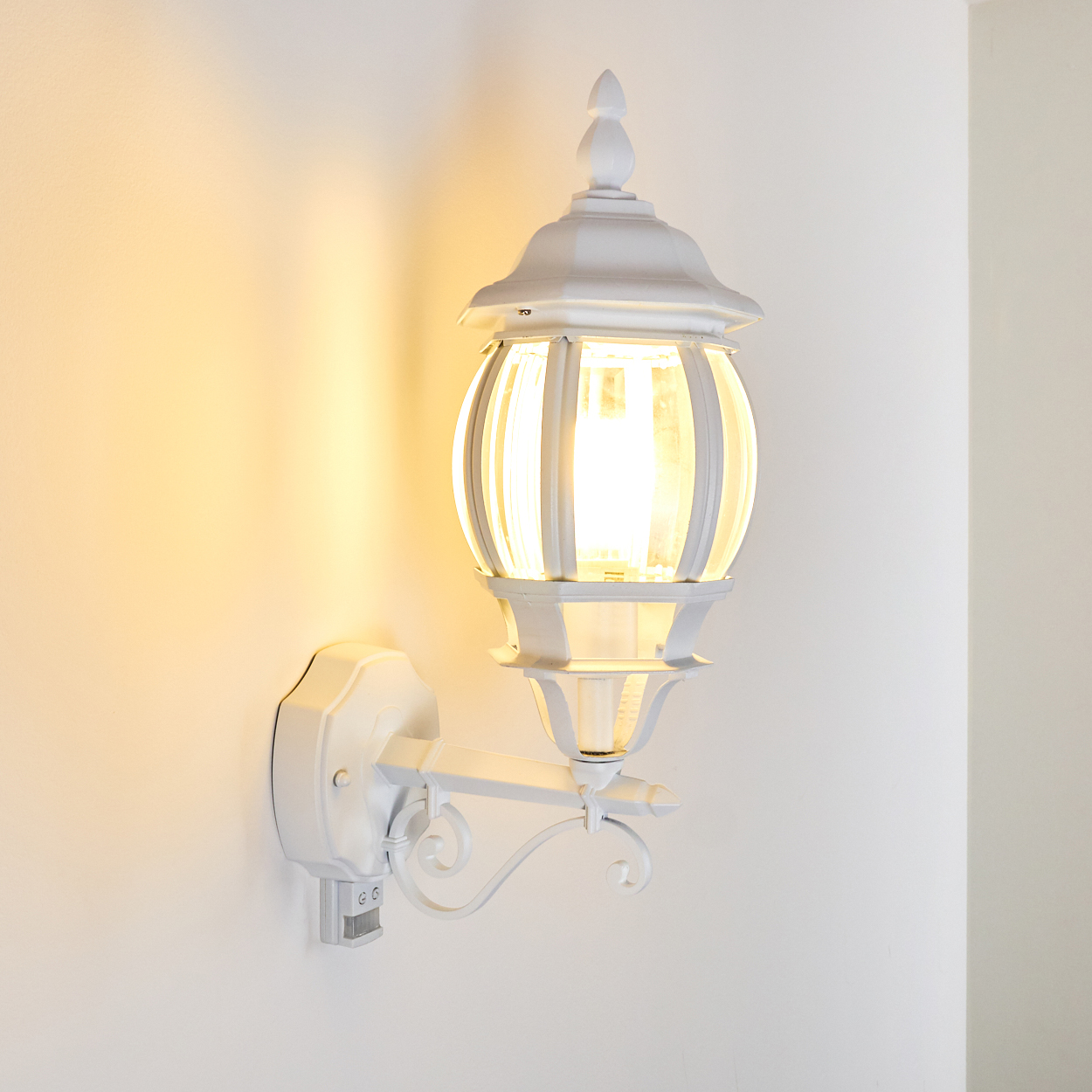 Eine weiße Außenlampe ist an einer weißen Wand montiert. Die Lampe ist eingeschaltet und das Licht leuchtet. Es hat ein klassisches Design.