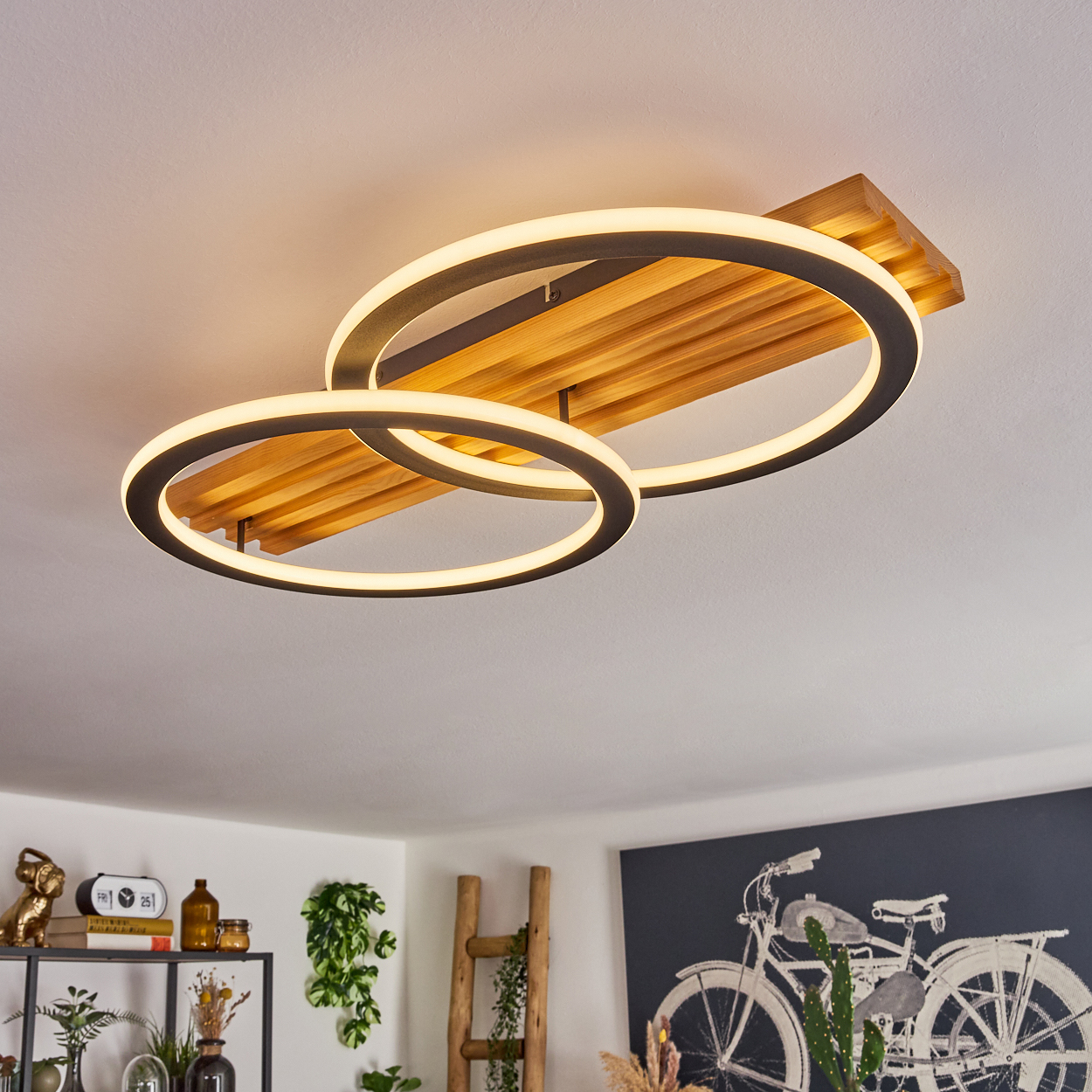 Deckenlampe mit zwei sich kreuzenden Lichtringen. Holzsockel mit horizontalen Lamellen. Weiße Decke. Kunstwerk und Deko im Hintergrund.